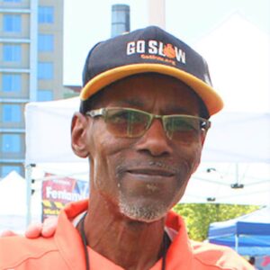 African-American man wearing an orange shirt and black and orange hat that reads "Go Slow" smiles. There is a building, white tent and blue tent in the background. 